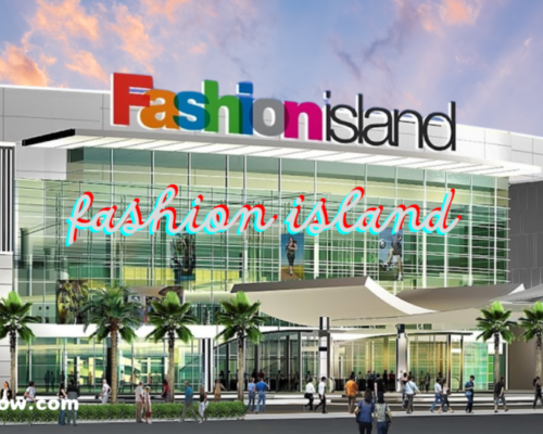 An exterior view of a shopping center named "Fashion Island," with a colorful logo at the top of the building. Below, there are glass windows, palm trees, and people walking around the entrance.